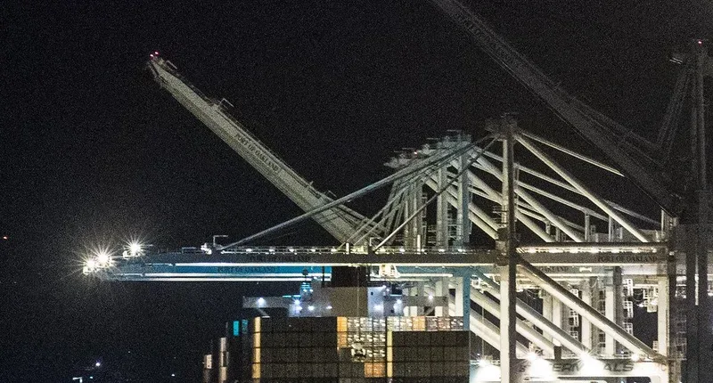 夜の港で、照明に照らされた大きなクレーンがコンテナ船の上に腕を伸ばしている写真